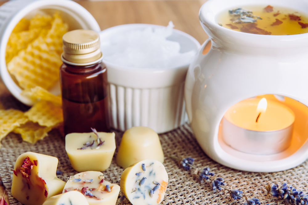 Using wax melts in aromatherapy lamp diffuser at home. Some melts ingredients including beeswax, solid coconut oil, essential oil, dried flowers shown on table arranged around tealight burner.