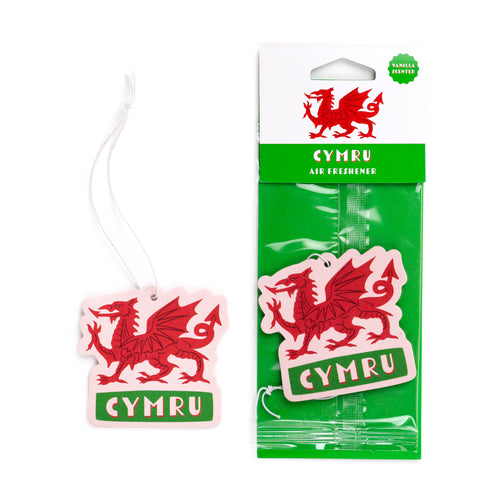 Front view of a vanilla Welsh Dragon air freshener and its packaging on a white background. Both items feature a red dragon on a light pink background with 'CYMRU' in green. The unpackaged air freshener hangs by a white string. The packaged air freshener is sealed in clear plastic with a green backing and a white card header displaying the red dragon, 'CYMRU AIR FRESHENER', and a 'VANILLA SCENTED' label.
