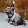 Angled view of an intricate wood-effect elephant backflow incense burner, with a lit incense cone on its back and smoke cascading down its side.
