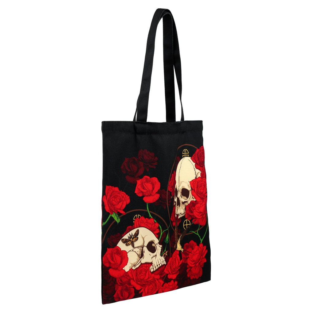 Angled black reusable shopping tote bag, featuring a design of pale skulls, one with a moth, amidst vibrant red roses.