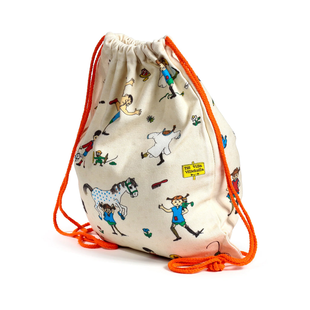 Angled view of a cream canvas drawstring bag, cinched at the top, displaying a colourful Pippi Longstocking pattern with characters including Pippi and a horse, plus a 'Till Villa Villekulla' sign, and bright orange drawstrings.