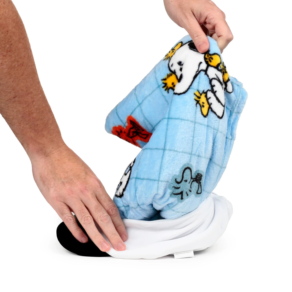Close-up: hands folding a blue, grid-patterned, soft-feel Snoopy and Woodstock blanket into the white plush toy, showing its black base and small fabric tag.