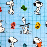 Close-up of a blue plush blanket featuring a grid pattern and multiple cartoon Peanuts Snoopy and Woodstock characters. Snoopy is shown jumping, wearing sunglasses, and holding a red heart, with Woodstock in green, orange, and yellow.