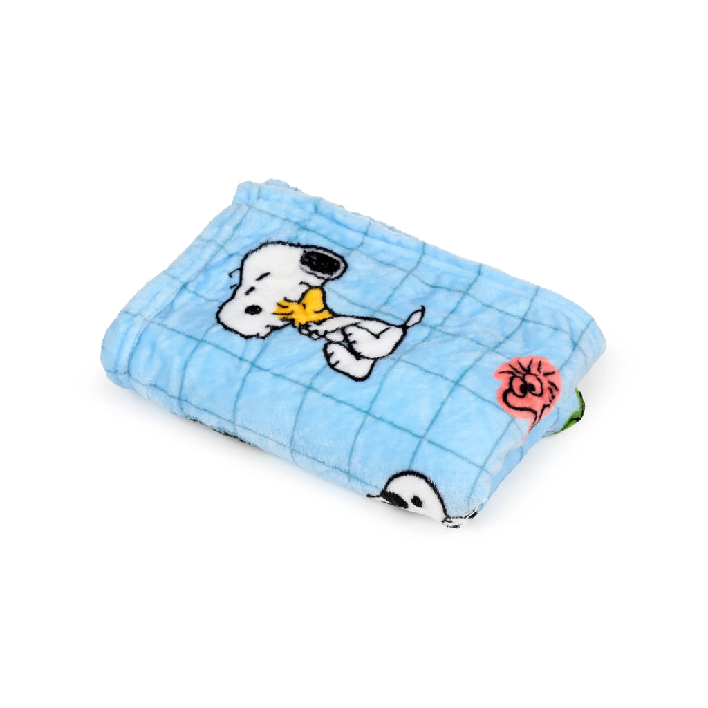 Angled view of a folded light blue plush Peanuts travel blanket, depicting Snoopy and Woodstock characters amidst a subtle grid pattern.