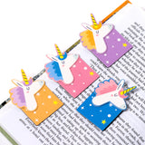 Angled close-up of four colourful magnetic unicorn bookmarks. Each bookmark features a white unicorn head with a striped yellow horn and a pastel-coloured mane (lilac, blue, pink, and orange), attached to a rectangular base patterned with stars and dots in corresponding or contrasting hues (lilac, pink, blue, and orange). The bookmarks are shown marking pages in an open book.