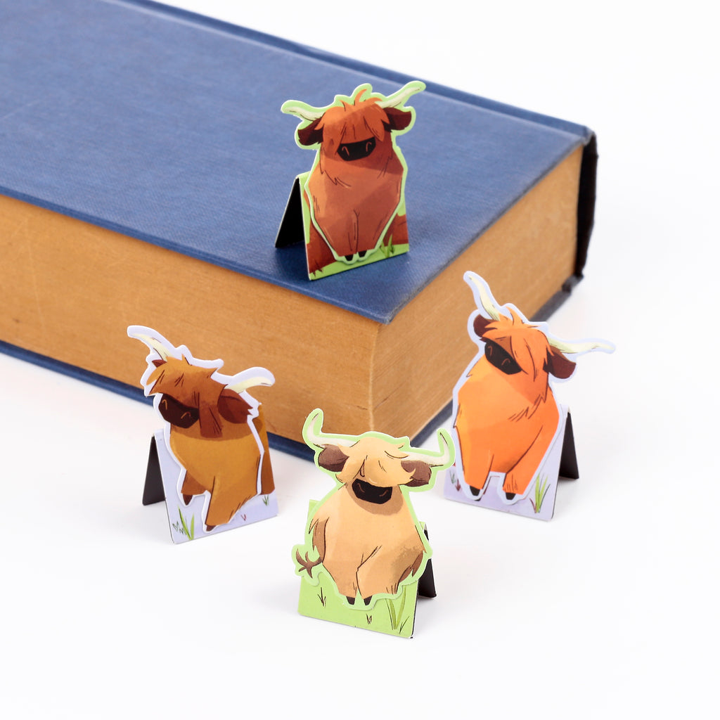 Angled view of four magnetic Hairy Coo bookmarks. One brown Highland Coo bookmark is shown positioned on a closed blue book, demonstrating its use, while three other cartoon coo illustrations in varying shades stand upright on a white background.