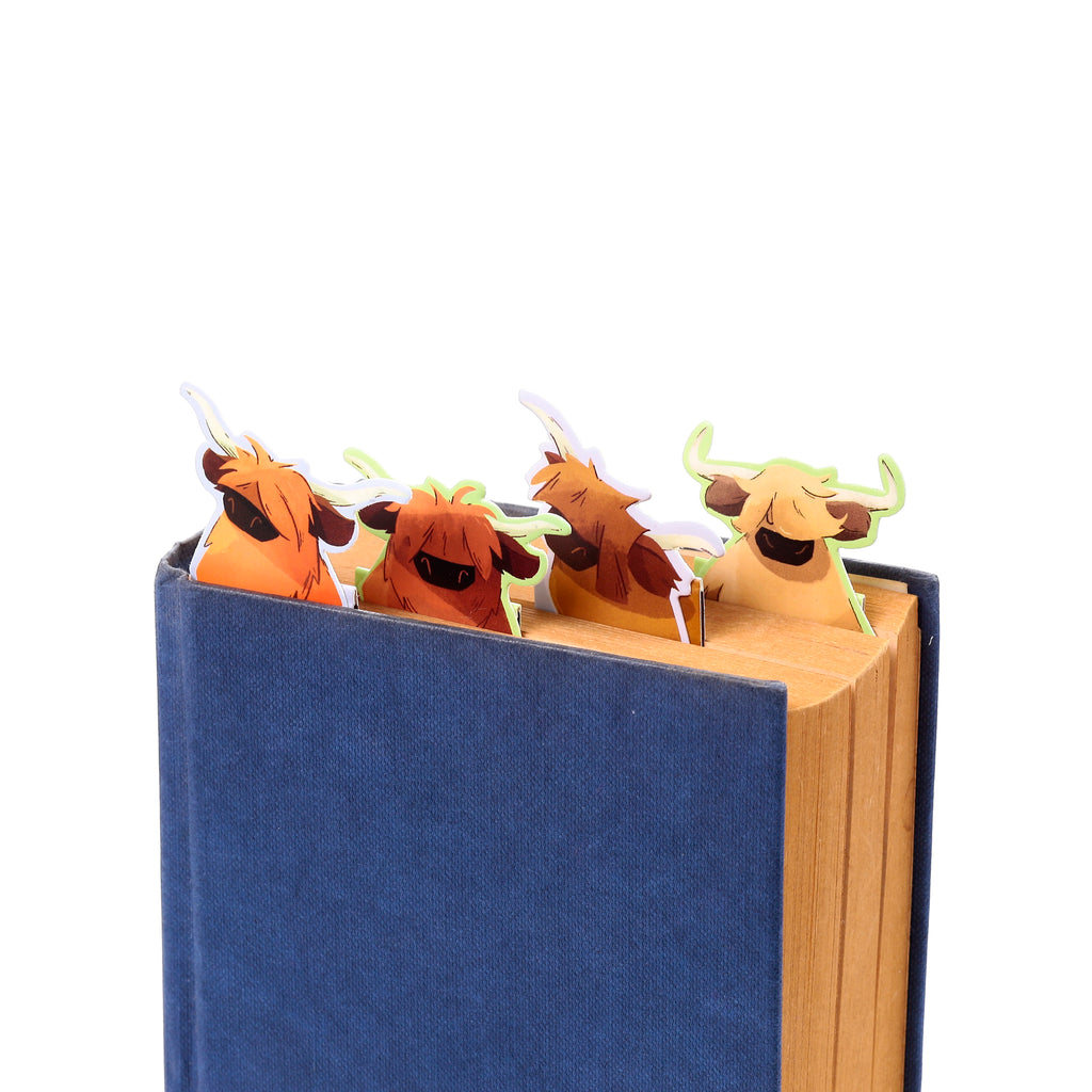 Close-up of four magnetic bookmarks depicting cartoon hairy Highland coos, in shades of brown and orange, peeping from a dark blue book.
