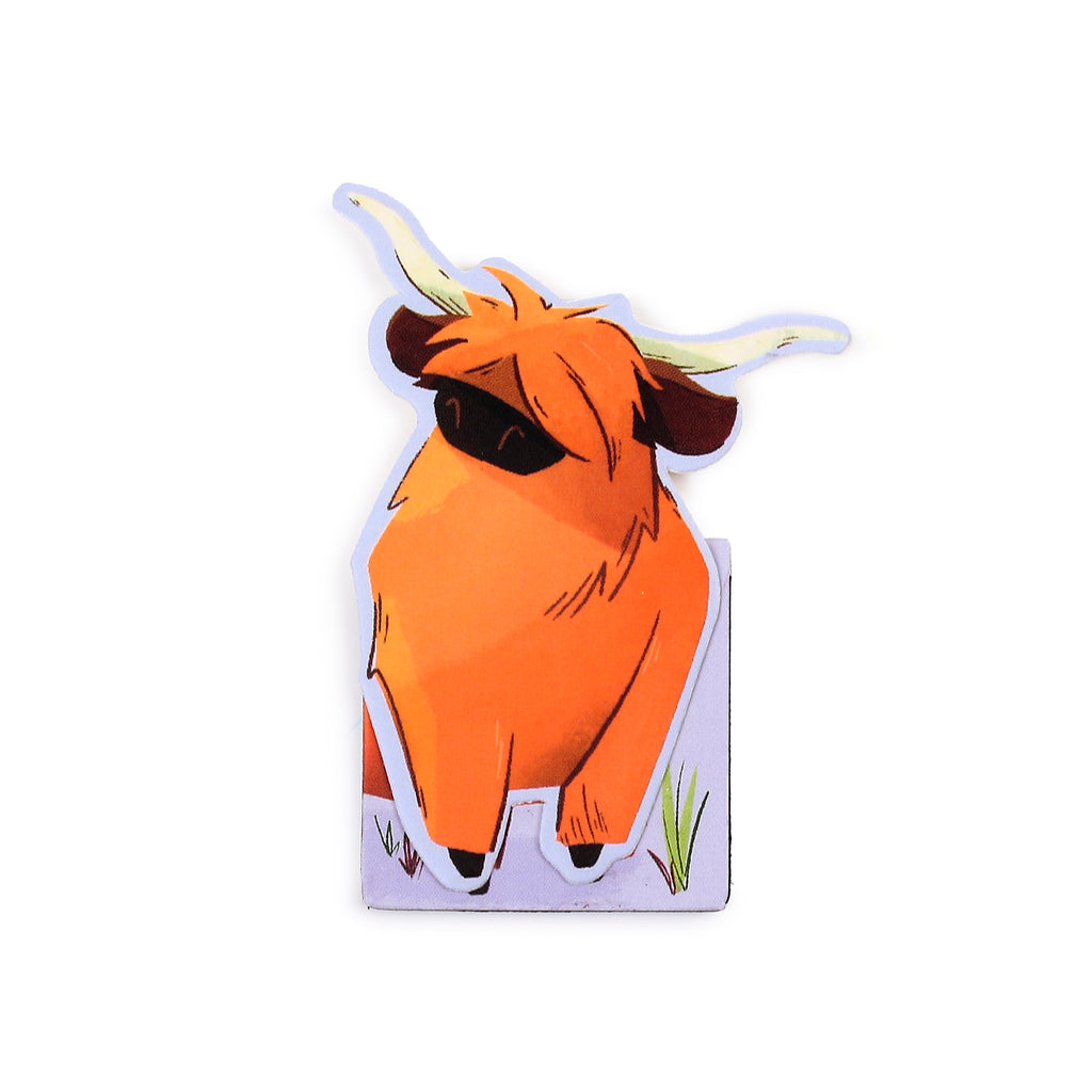 Front view of a magnetic bookmark shaped like an orange Hairy Coo. The Highland cattle illustration shows shaggy hair covering its eyes, horns, and a small patch of grass at its base, all set against a white background.