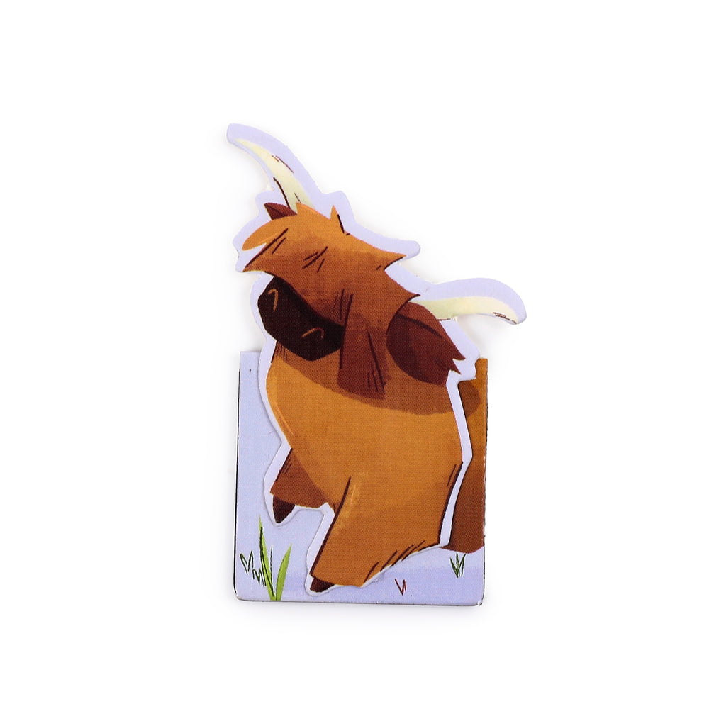 Close-up of a magnetic bookmark depicting a cartoon Highland coo with shaggy brown fur and light horns, standing on a light blue and white base with green grass details.