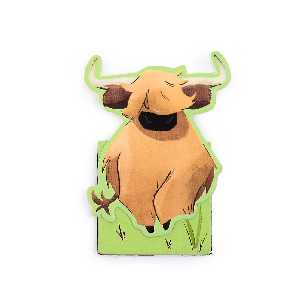 Close-up of a magnetic bookmark depicting a cartoon Highland Coo with shaggy light brown fur, brown horns, and a green grass base.