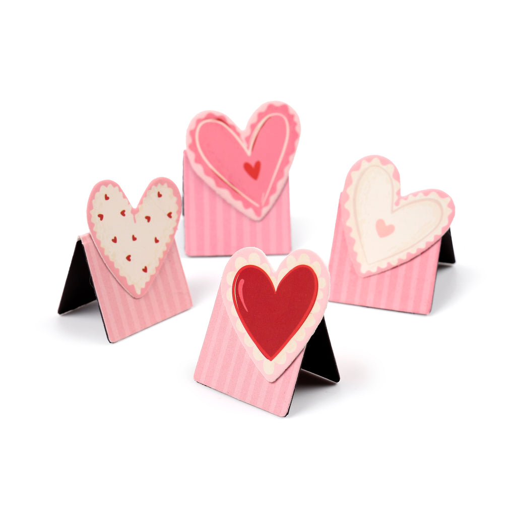 Angled view of four pink and white magnetic bookmarks, each featuring heart designs and striped patterns, standing upright on a white background.