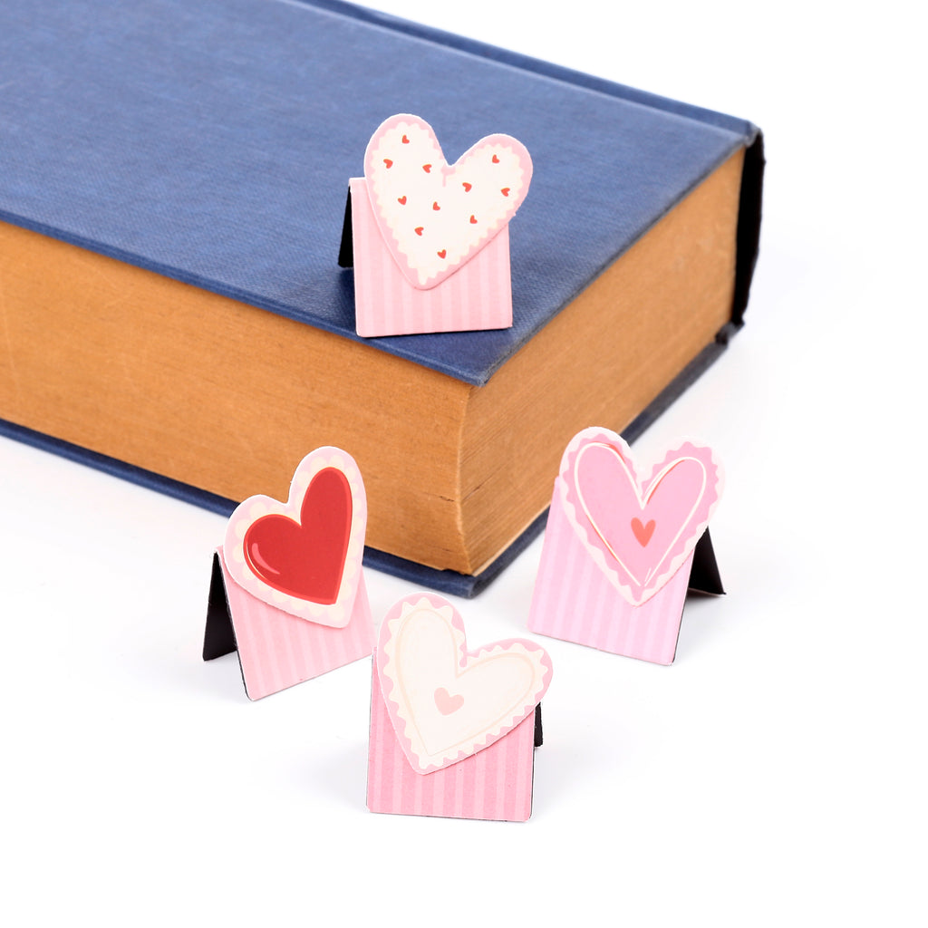 Angled view of four pink and red heart-themed magnetic bookmarks, with one placed on a blue book and three others arranged on a white background.