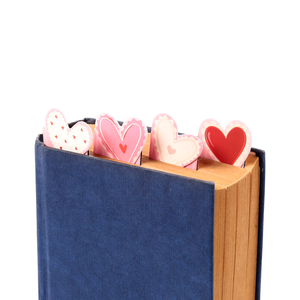 Angled close-up of Amore Heart Magnetic Bookmark Set, featuring four pink, white, and red heart-shaped bookmarks, some with patterns, marking pages in a blue book.