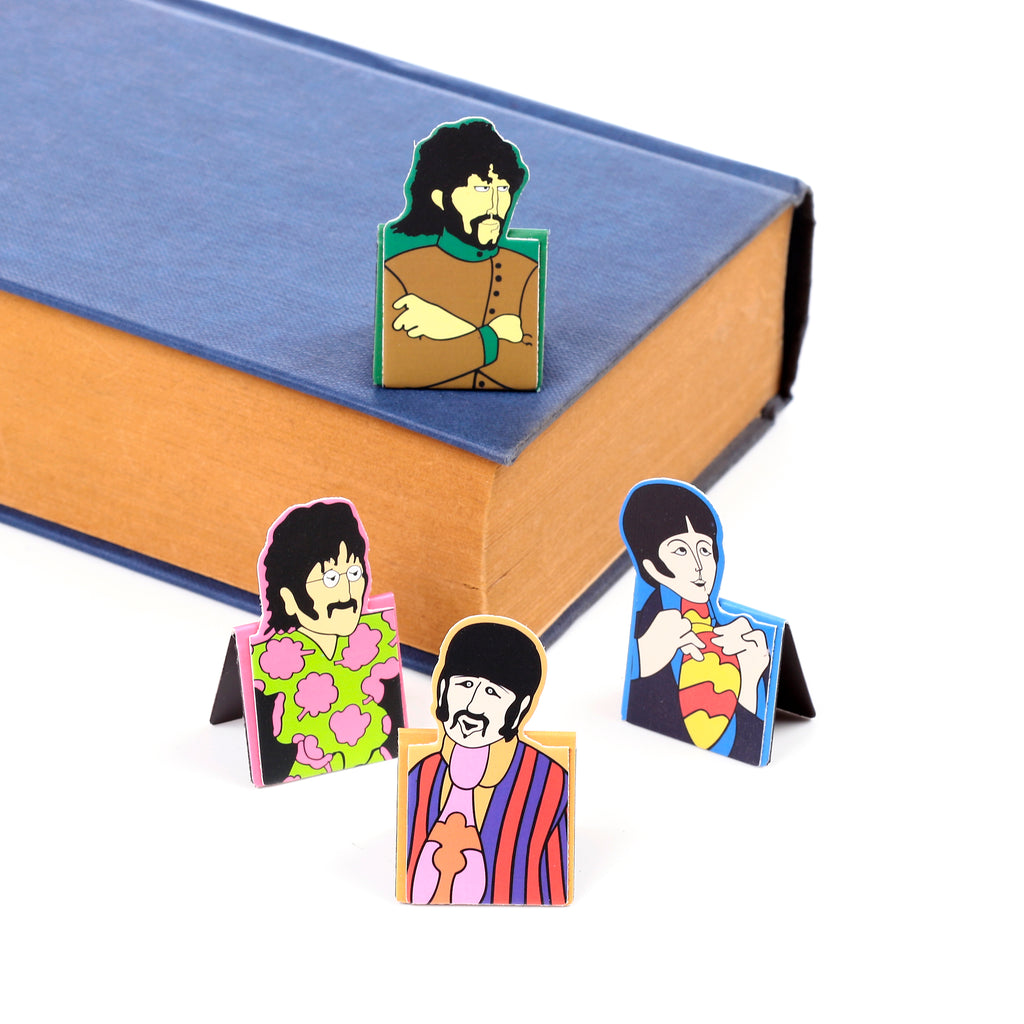 Angled view of four colourful magnetic bookmarks, each featuring a stylised The Beatles Yellow Submarine character, displayed with one bookmark marking a page in a blue hardback book.