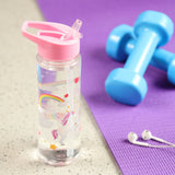 Angled lifestyle shot of a clear reusable water bottle with a pink flip straw, featuring an enchanted unicorn and colourful rainbow design, with KEEP GOING TO THE END OF THE RAINBOW text, filled with clear water. Blue dumbbells and white earphones rest on a purple exercise mat in the blurred background.