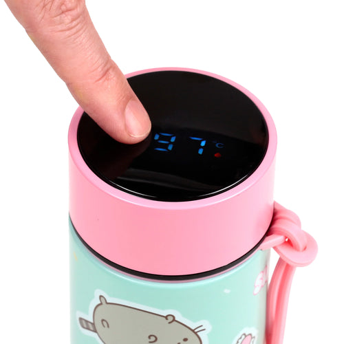 Close-up angled view of a reusable insulated drinks bottle, showing a finger pressing the digital thermometer display on its pink lid, which reads 97°C. The mint green body features a cat and sweet pattern.