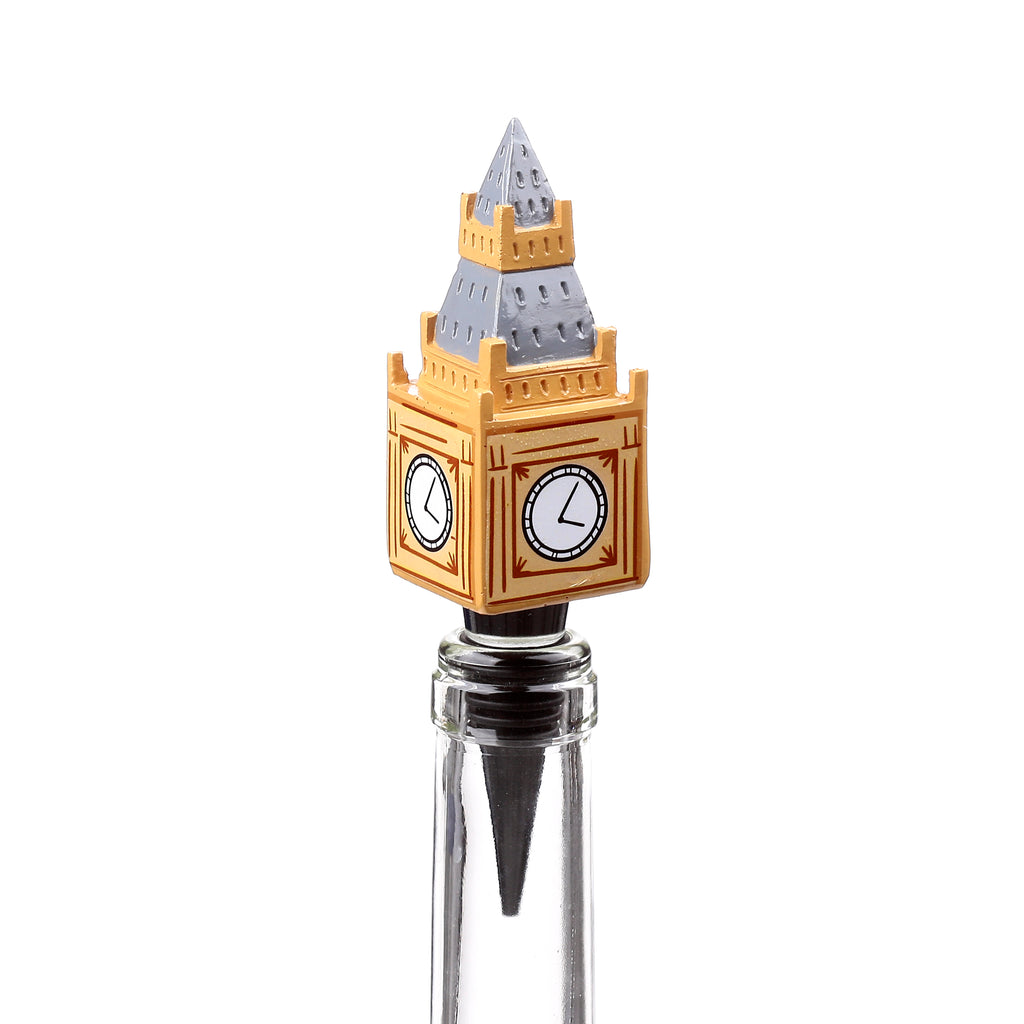 Novelty bottle stopper shaped like the Big Ben clock tower, with golden-coloured walls, grey spire, and two visible clock faces, inserted into a clear glass bottle neck, from an angled view.