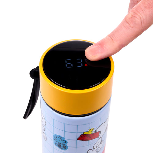 Close-up of a finger pressing the digital thermometer display on a Peanuts Snoopy & Woodstock insulated flask, showing 63°C. The flask features a yellow cap and a light blue body with character illustrations.