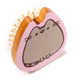 Angled view of a bamboo hair brush shaped like Pusheen the Cat, featuring a grey cat face with a pink outline and light-coloured wooden pins.