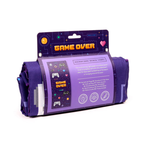Angled view of a rolled purple microfibre beach towel, visible in its packaging. The packaging features a vibrant pixelated 'Game Over' design with orange text, gaming controllers, hearts, and coins, alongside text detailing its quick-drying and ultra-absorbent qualities.