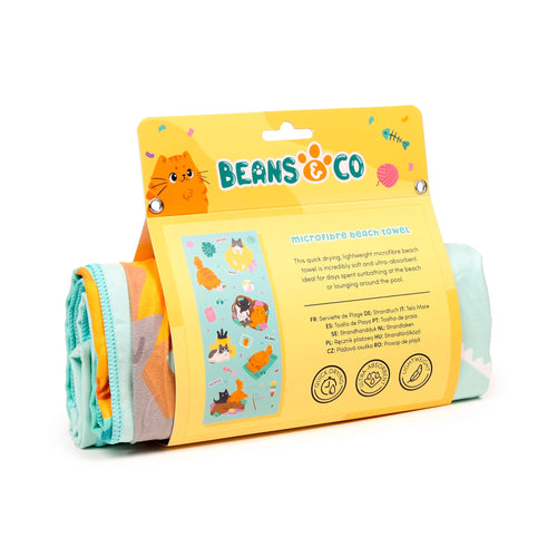 Angled view of a rolled microfibre beach towel in yellow retail packaging. The packaging displays cartoon cat illustrations and text highlighting the towel's quick-drying and ultra-absorbent properties. Visible sections of the towel are turquoise, orange, and grey.