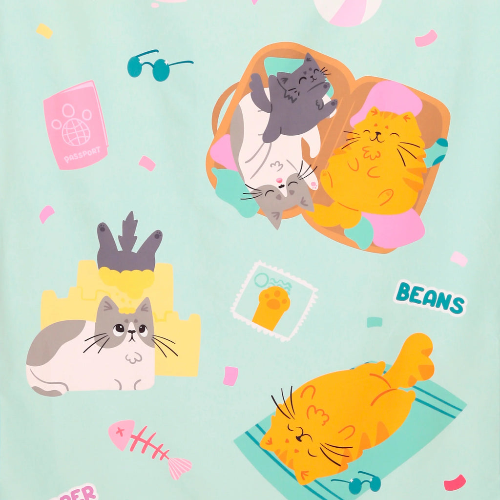 Close-up of a beach towel featuring a fun, colourful illustration on a mint green background. The design shows cartoon cats enjoying a holiday, with some in an open suitcase, one by a yellow sandcastle, and another sunbathing with sunglasses. Other details include a pink passport, a fish skeleton, and the word 'BEANS'.