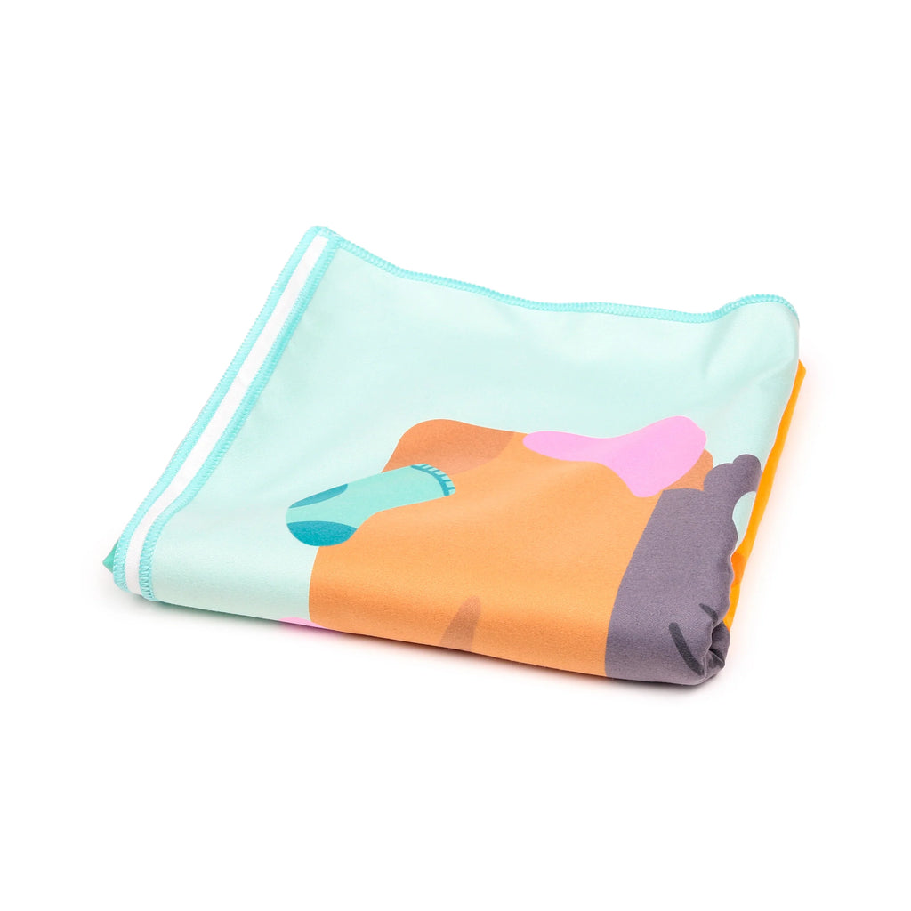 Folded microfibre beach towel with a colourful abstract cat design, featuring light blue, orange, pink, and dark grey patterns, shown from an angled view.