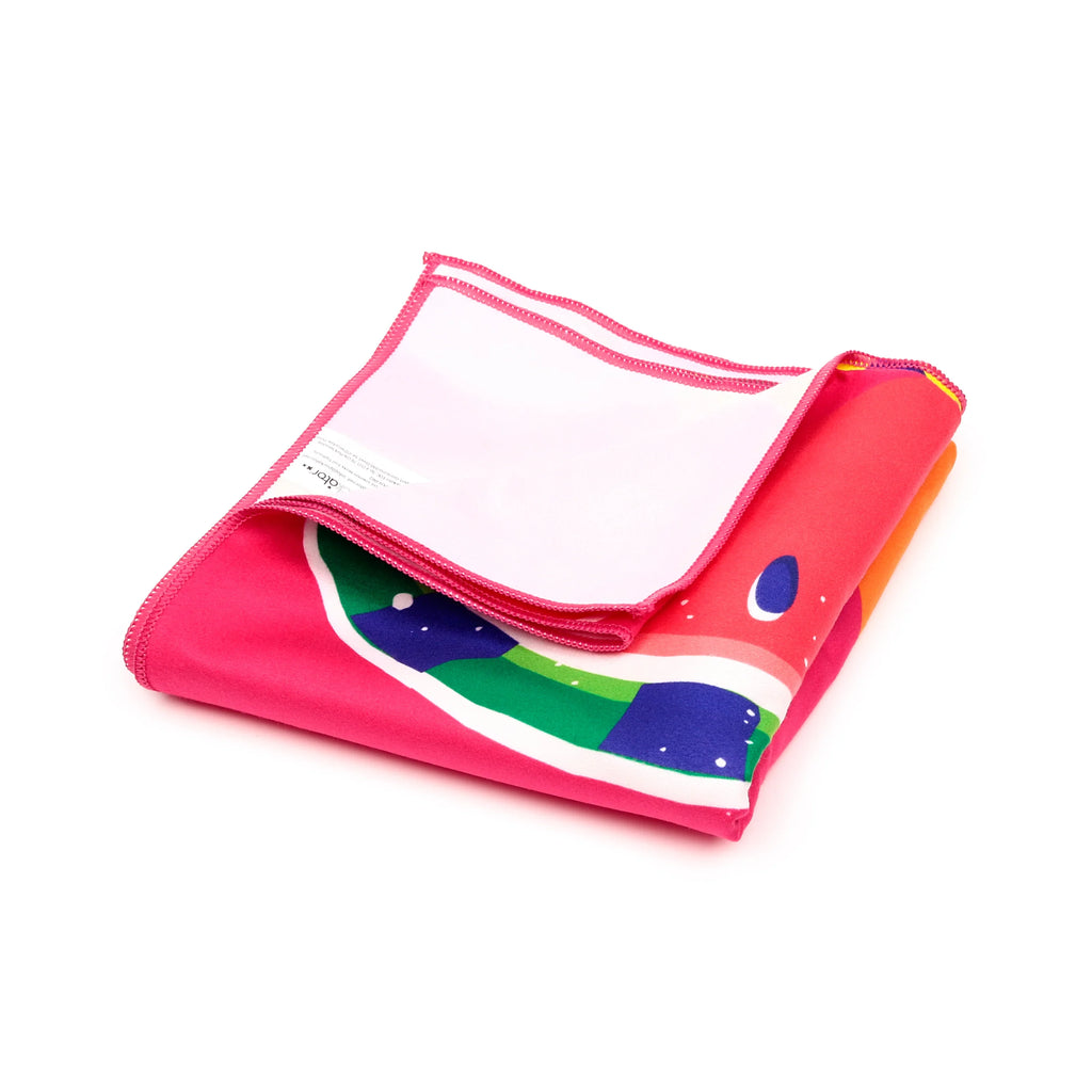 Angled view of a folded vibrant pink microfibre beach towel with overlocked edges and a colourful summer fruits pattern featuring green and blue elements, on a white background.