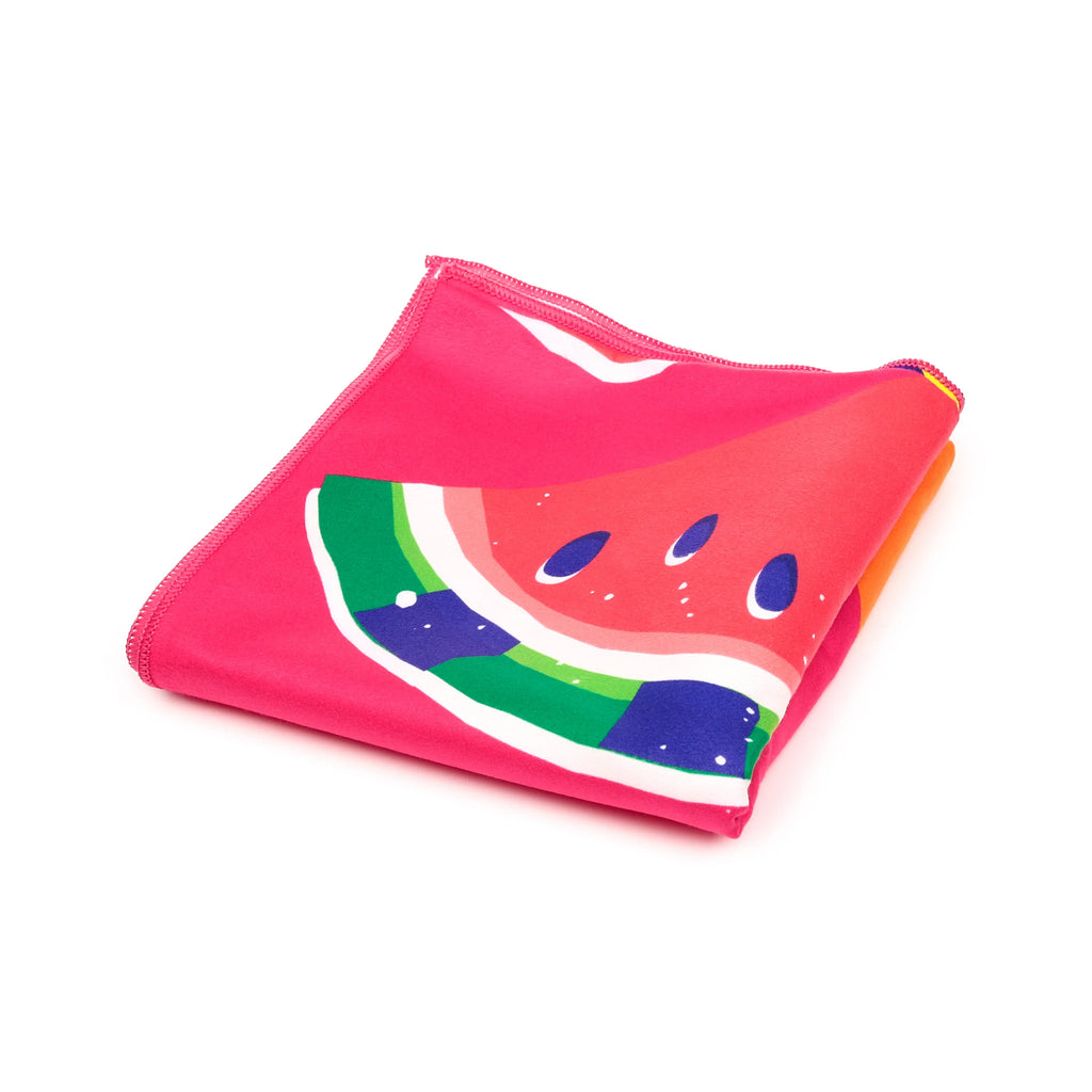 Angled view of a folded, bright pink microfibre beach towel with a vibrant watermelon slice pattern featuring red, green, white, and blue seeds.