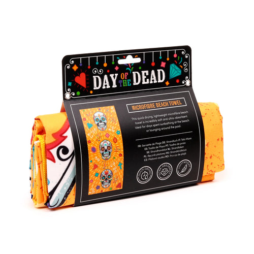 Angled view of a rolled orange microfibre beach towel in its black packaging sleeve. The packaging prominently displays a colourful Day of the Dead sugar skull pattern and text, plus icons for quick-drying, ultra-absorbent, and lightweight features.