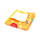 Angled view of a folded microfibre beach towel, yellow-orange with abstract colourful patterns in red, green, blue, and purple, a speckled texture, and yellow stitched edges. A white label is visible on the top fold.