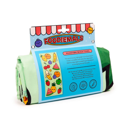 Rolled-up light green microfibre beach towel in its bright blue retail packaging, featuring 'Foodiemals' cartoon watermelon and other summer fruit designs.
