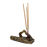 Angled view of a metallic gold and bronze Buddha hand incense holder, featuring intricate henna-style patterns, with three light brown incense sticks, with purple bases, inserted into the palm, on a white background.