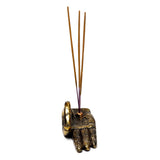 Angled view of a gold-coloured, hand-shaped incense holder, intricately decorated with henna-style patterns across its palm and fingers, featuring three brown incense sticks, against a white background.