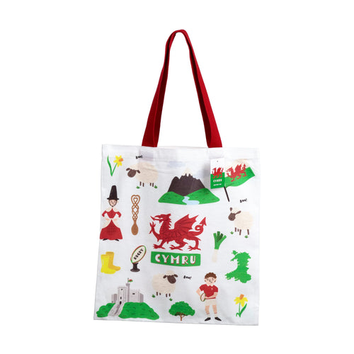 Front view of a white shopping tote bag with red handles, featuring a vibrant Welsh illustration design including a red dragon, 'CYMRU' text, sheep, daffodils, and a castle.