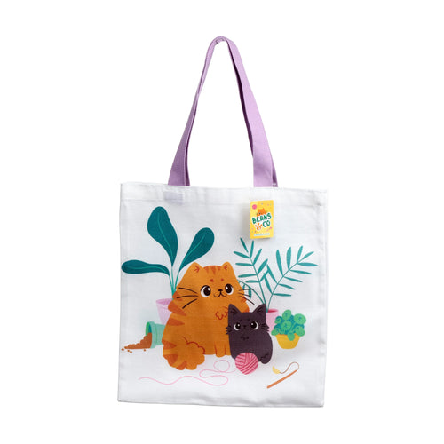 Front view of a white shopping tote bag with lilac handles, featuring a colourful illustration of a ginger cat and a black cat sitting amongst potted green houseplants, a pink ball of yarn, and spilt brown soil.