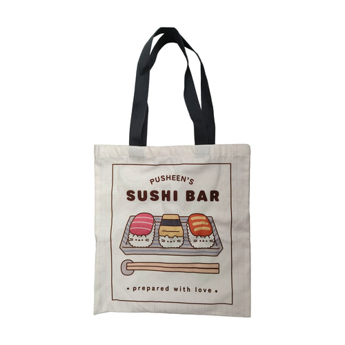 Front view of an off-white shopping tote bag with black handles, featuring a colourful 'Pusheen the Cat Sushi Bar' design. The design includes three sushi pieces with Pusheen's face, chopsticks, and 'prepared with love' text.