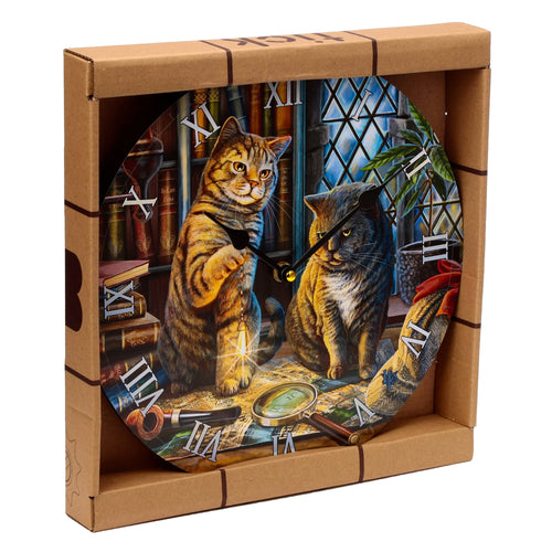 Angled view of a decorative round wall clock in its brown cardboard packaging, depicting two cats in a library scene with books, a map, a magnifying glass, and a pipe. The clock features black hands and Roman numeral hour markers.