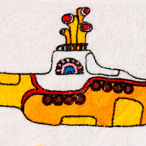 Close-up of a white travel towel showing The Beatles Yellow Submarine design. The yellow submarine features red and blue details, brown porthole accents, and yellow upper structures on its textured surface.