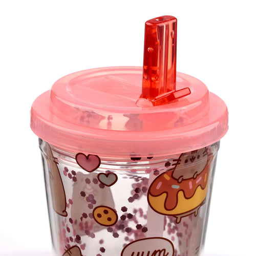 Close-up angled view of a clear double-walled cup with a pink lid and red straw, showcasing a Pusheen foodie design with a doughnut, cookie, hearts, and floating pink hexagonal glitter.