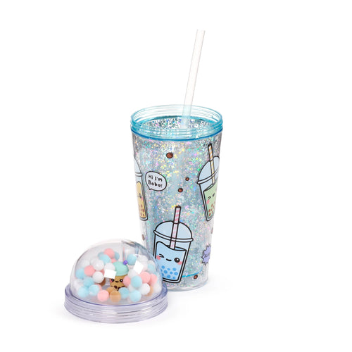Angled view of a double-walled clear plastic cup with a light blue lid and a white straw. The cup features iridescent glitter, star confetti, and cartoon bubble tea illustrations. Next to it, a clear dome charm contains pastel pom-poms and a small cartoon figure.