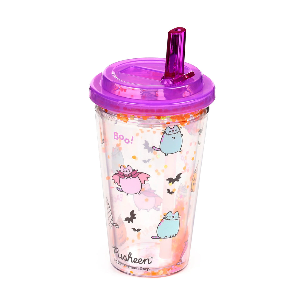 Angled view of a clear, double-walled cup with a purple lid and straw. The cup features Halloween Pusheen the Cat designs, including a vampire cat, stitched cats, bats, and 'Boo!' text, with loose orange and pink glitter between the walls.