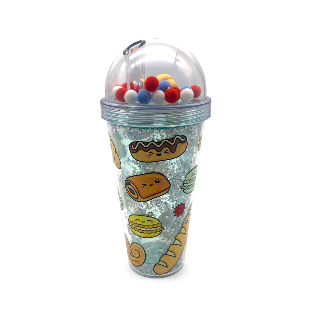 Front view of a clear double-walled cup with a dome lid, containing red, white, and blue pom-pom charms and a straw. The cup features glitter with stars and cartoon French bakery illustrations.