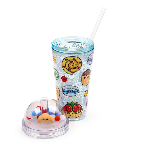 Angled view of a clear double-walled cup with a straw, featuring a cartoon baked goods design and internal glitter. A clear dome lid with a croissant charm and colourful pom-poms sits beside it.