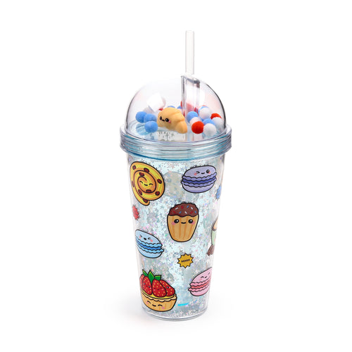 Angled view of a Foodiemals Boulangerie double-walled cup with a clear straw. The cup's outer wall features cartoon dessert illustrations including cupcakes, macarons, a strawberry tart, and a swirled pastry, set against a sparkly inner wall filled with star-shaped glitter. The clear dome lid contains a smiling croissant charm alongside red, white, and blue fluffy pom-poms.