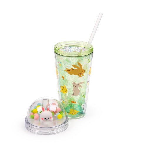 Angled view of a double-walled cup with a green glittery interior and spring-themed illustrations of bunnies, chicks, and carrots. A white straw protrudes from the cup. Beside it, the clear lid features a dome containing a white rabbit charm amongst colourful pom-poms.