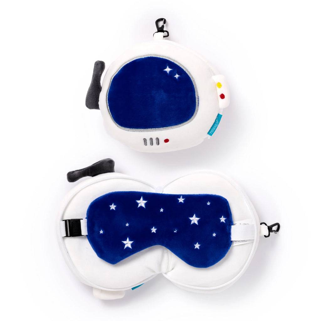 Overhead view of a plush white 'Space Cadet' travel pillow and eye mask set, displayed both packed and unpacked. The packed round pillow, shaped like an astronaut's helmet, features a deep blue visor with white embroidered stars, red and yellow details, and a black carabiner clip. The unpacked pillow reveals a matching deep blue eye mask with white stars and an adjustable strap.