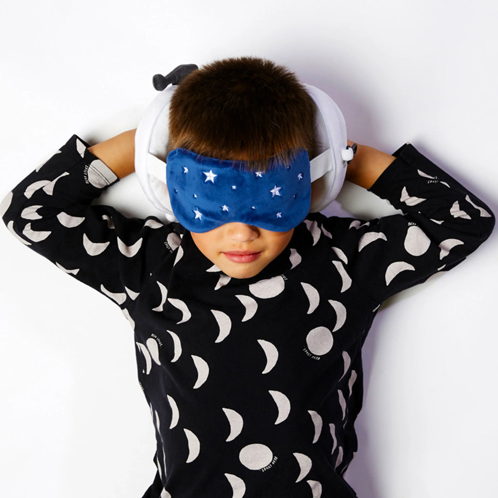 Lifestyle shot of a child lying down, wearing a blue plush eye mask with white star details and resting on a white plush round travel pillow.