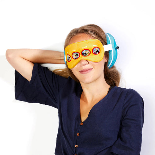 In-context lifestyle shot of a smiling person wearing a yellow travel eye mask adorned with circular illustrations of John, Paul, George, and Ringo. A turquoise plush round travel pillow is visible behind their head.