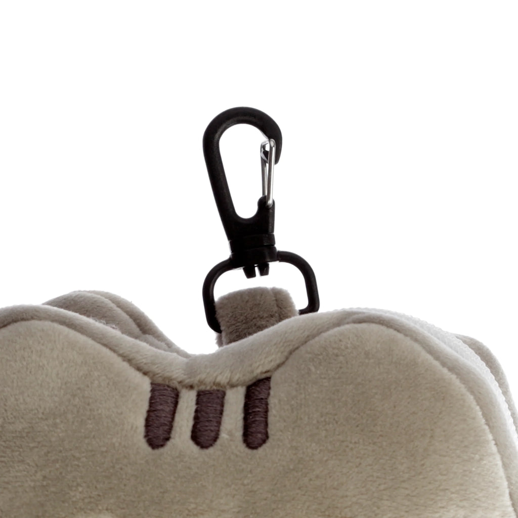 Close-up of a black carabiner clip attached to a light beige plush travel pillow, featuring dark brown embroidered stripes.